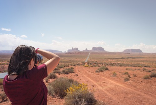 A person takes a photo of a desert landscape with distant rock formations under a clear sky.