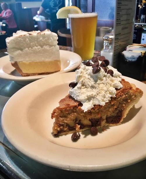 A slice of dessert topped with whipped cream and chocolate chips, next to a large piece of cream pie and a beer.