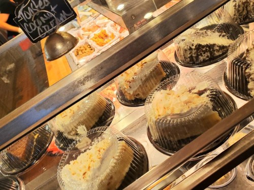 Display of various cakes in clear containers at a bakery, with a chalkboard sign in the background.