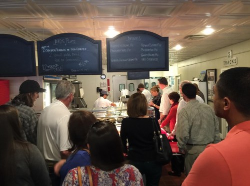 A busy cafeteria line with people waiting to order food, featuring a menu board and staff serving dishes.