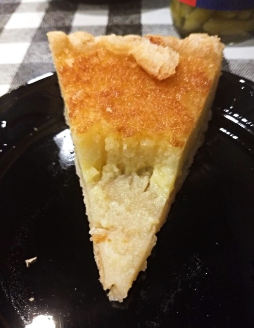 A slice of golden, baked dessert on a black plate, with a flaky top and a moist, creamy interior.