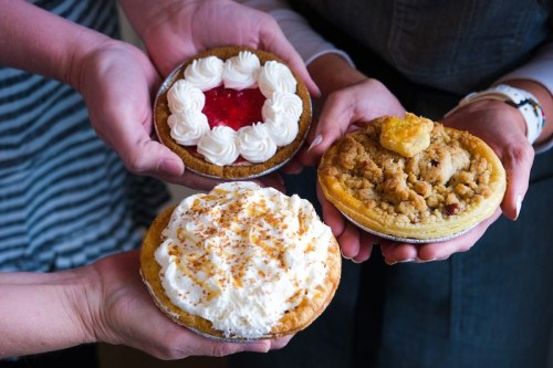 Three hands hold different mini pies: one with red topping, one with whipped cream, and one with a crumbly topping.