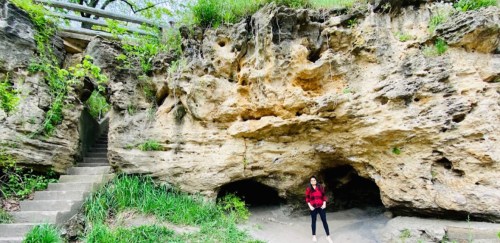 A person stands in front of a large rock formation with a cave entrance, surrounded by greenery and steps nearby.