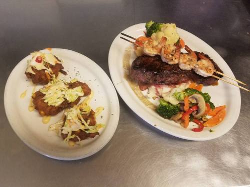 A plate with grilled shrimp skewers and steak, accompanied by sautéed vegetables and two fried dishes topped with sauce.