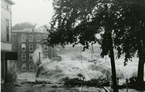 A historic black-and-white photo of a flooded street, with water rushing past buildings and trees during a storm.