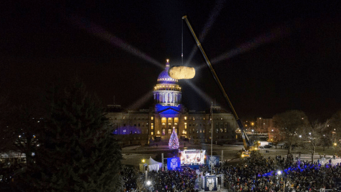 A large potato is lifted by a crane above a crowd at night, with a lit building and Christmas tree in the background.