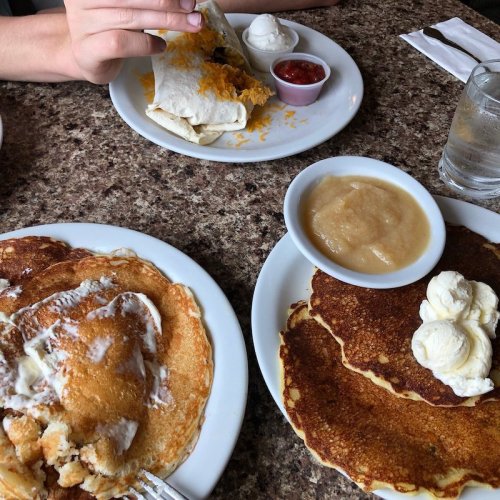 A table with pancakes, a plate of enchiladas, applesauce, and a small bowl of sour cream, with a hand reaching for food.