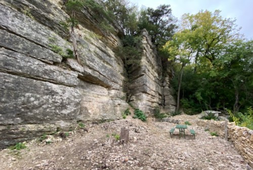Rocky cliffside with layered stone formations, surrounded by trees and a small clearing with a picnic table.