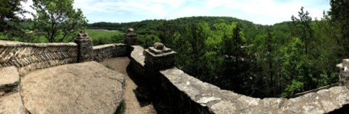 A panoramic view of a stone railing overlooking lush green hills and trees under a clear blue sky.