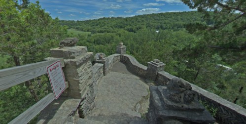 A stone overlook with a railing, surrounded by lush green trees and a blue sky with clouds in the background.