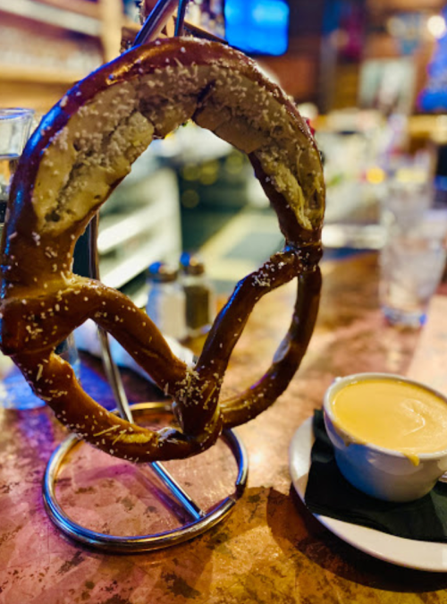 A large pretzel on a stand, sprinkled with salt, next to a small bowl of cheese dip on a bar table.