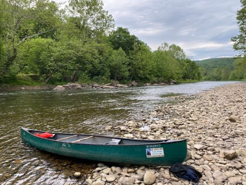 A green canoe rests on a rocky riverbank surrounded by lush greenery and a calm river under a cloudy sky.