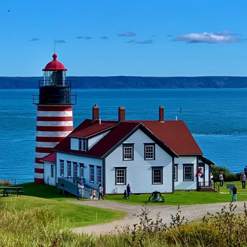 A red and white striped lighthouse beside a white house, overlooking a calm blue sea with people enjoying the scenery.