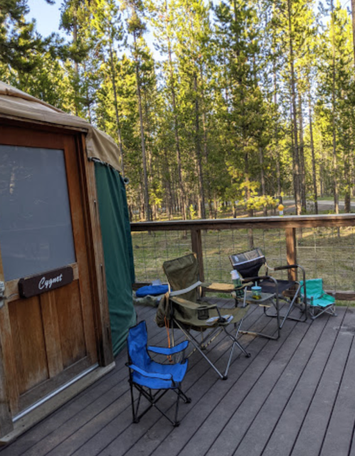 A wooden deck with camping chairs surrounded by tall trees and a yurt entrance in the background.