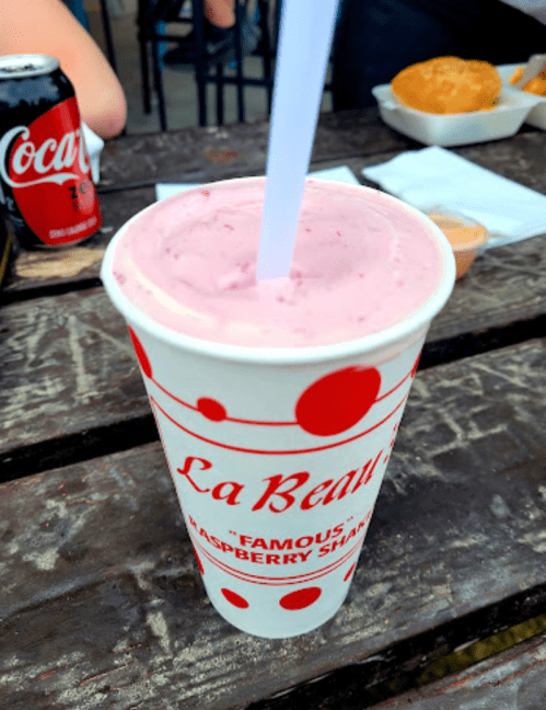 A cup of pink raspberry shake with a straw, placed on a wooden table next to a can of Coke and food.