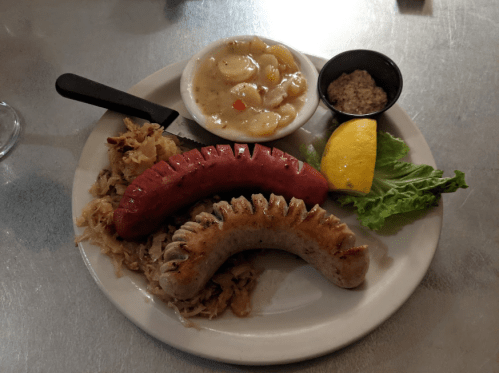 A plate with two types of sausage, sauerkraut, a lemon wedge, lettuce, and a small bowl of mustard and pickles.