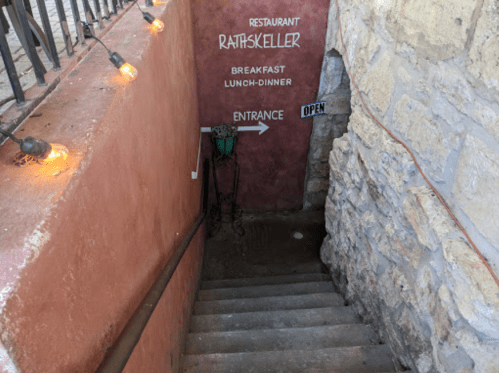 Stairs leading down to a restaurant entrance, with a sign indicating "Rathskeller" and "Open." Warm lights above.