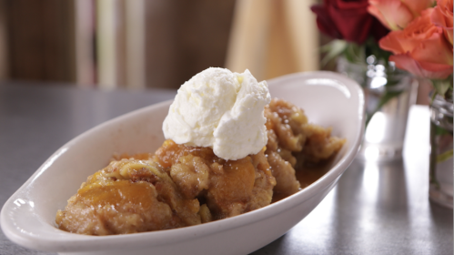 A dish of peach cobbler topped with a scoop of whipped cream, served in a white bowl with flowers in the background.