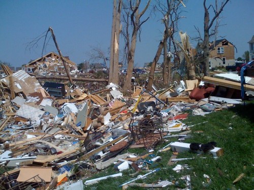 Debris and destruction from a tornado, with damaged trees and a collapsed house in the background.