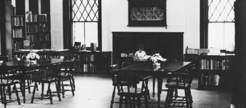A quiet library interior with wooden tables, chairs, and a person reading in the background.