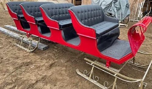 A red and black vintage sleigh with plush seating, resting on metal runners in a rustic outdoor setting.