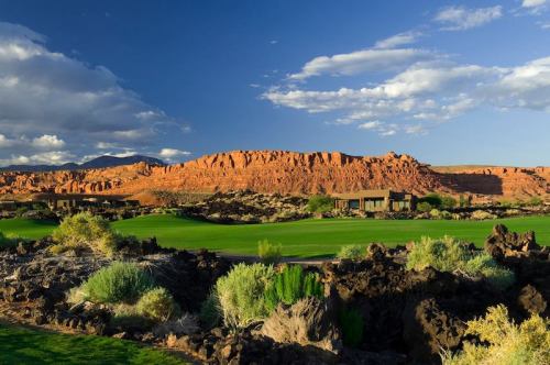 A scenic view of a golf course with lush green grass, surrounded by red rock formations and blue skies.