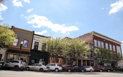 A row of historic buildings with trees lining the street under a blue sky with fluffy clouds.