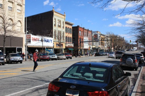 A street scene with historic buildings, parked cars, and a person crossing the road under a blue sky with clouds.