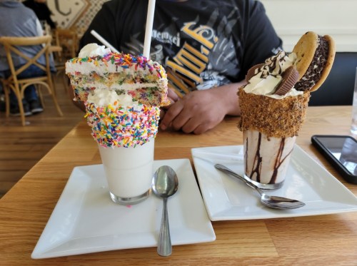 Two extravagant milkshakes on white plates: one topped with sprinkles and cake, the other with cookies and chocolate drizzle.