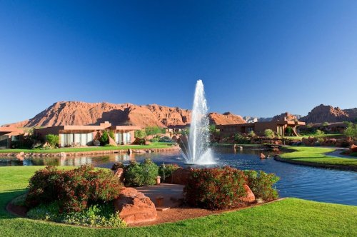 A serene landscape featuring a fountain in a pond, surrounded by lush greenery and red rock formations under a clear blue sky.
