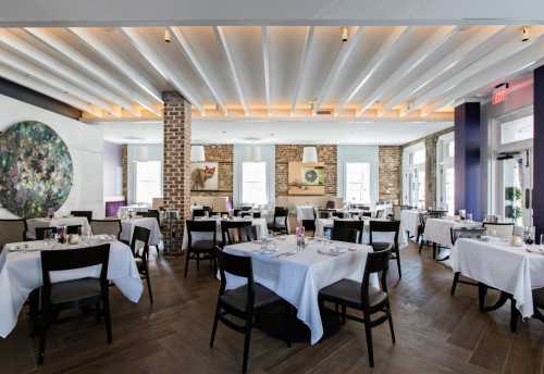 Elegant restaurant interior with white tablecloths, wooden floors, and modern decor, featuring a floral wall art piece.