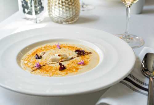 A bowl of creamy soup garnished with edible flowers and spices, served on a white tablecloth with a glass of wine.