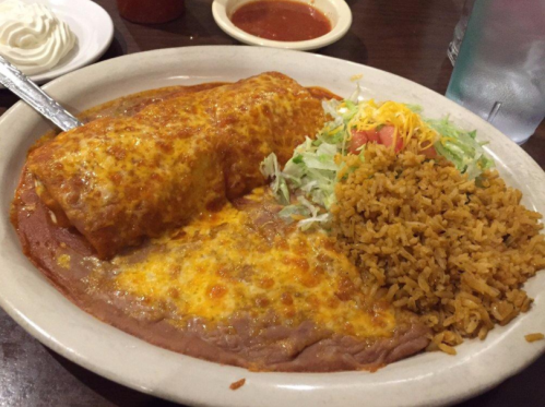 A plate of enchiladas topped with cheese, served with refried beans, rice, lettuce, and tomato.