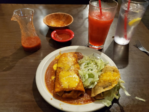 A plate of enchiladas topped with cheese, served with lettuce, a side of salsa, and drinks on a wooden table.