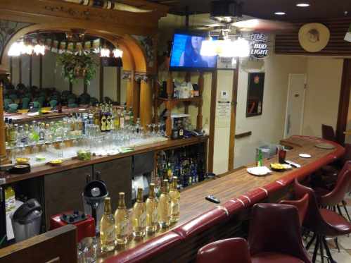 A bar with a wooden counter, shelves of liquor, glasses, and a TV in a cozy setting. Snacks are on the counter.