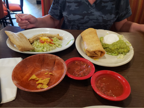 A person holds a fork near a plate with tamales, guacamole, and a taco, accompanied by salsa and tortilla chips.