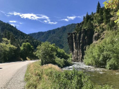 A scenic view of a river alongside a gravel road, with lush greenery and rocky cliffs under a blue sky.