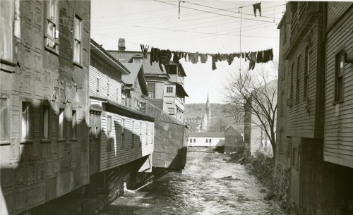 Black and white photo of a narrow street with buildings, a river, and laundry hanging between them.
