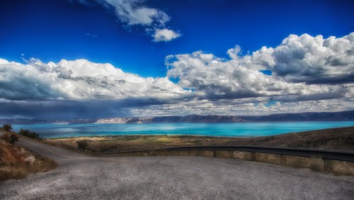A scenic view of a turquoise lake surrounded by mountains and dramatic clouds under a bright blue sky.