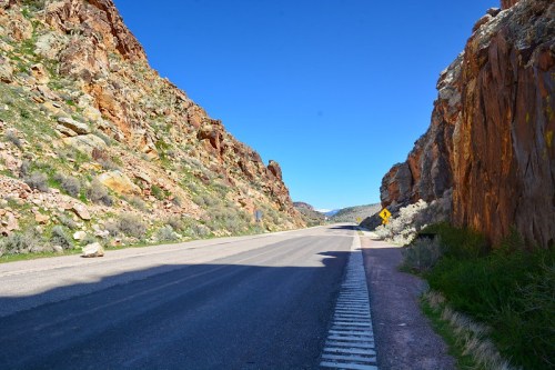 A scenic road flanked by rocky cliffs under a clear blue sky, with greenery along the roadside.