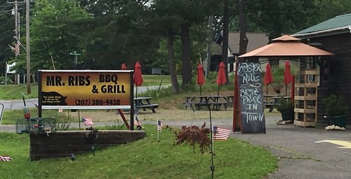 Sign for Mr. Ribs BBQ & Grill with outdoor seating and umbrellas, featuring a chalkboard with specials and American flags.