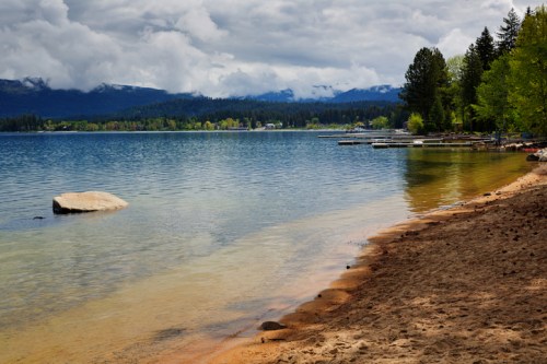 A serene lakeside view with sandy shore, calm water, and mountains under a cloudy sky. Green trees line the shore.