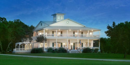 A large white house with multiple balconies and a porch, surrounded by greenery, illuminated at dusk.