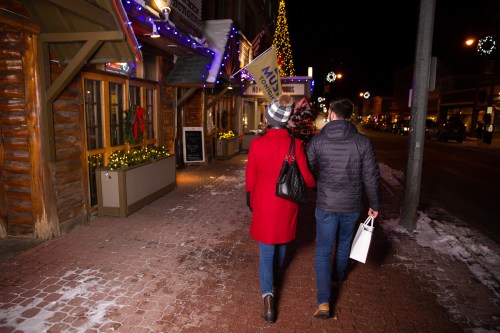 A couple walks hand in hand down a festive, snow-covered street, surrounded by holiday lights and decorations.