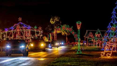 Colorful holiday lights decorate houses and trees along a dark road, with cars driving through the festive scene.