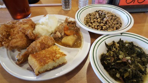 A plate of fried chicken, mashed potatoes, cornbread, black-eyed peas, and collard greens on a wooden table.