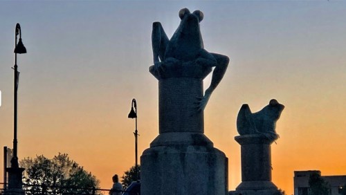 Silhouettes of frog sculptures on pillars against a sunset sky, with street lamps in the background.