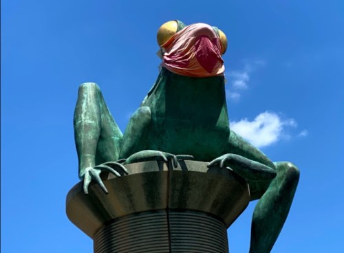 A large green frog statue wearing a pink mask, perched on a cylindrical pedestal against a blue sky.