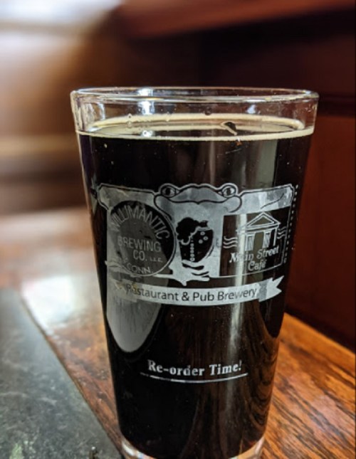 A clear glass of dark beer with a logo from a brewing company, placed on a wooden table.