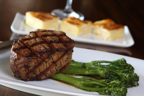 A grilled steak on a plate with broccolini and creamy potato gratin in the background.